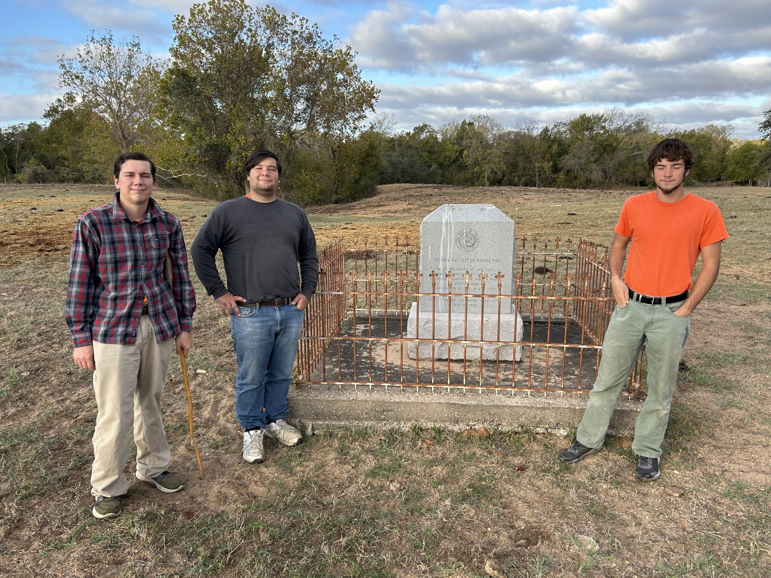 Three family members standing beside the fenced monument at the Woods Fort memorial site